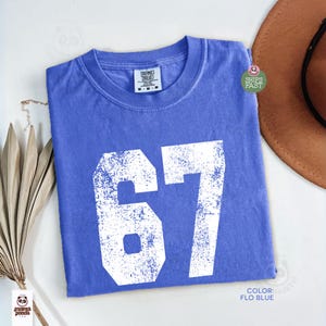 May include: A blue t-shirt with the number 67 in white, featuring a distressed, vintage design. The shirt is a solid color, with a crew neck and short sleeves. The color is described as "Flo Blue".
