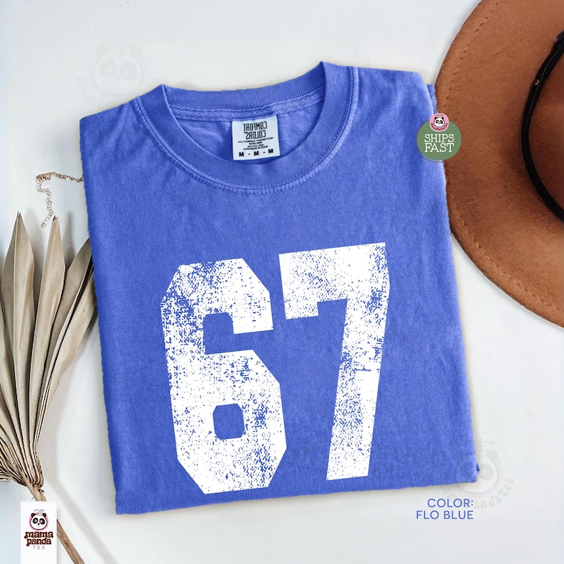 May include: A blue t-shirt with the number 67 in white, featuring a distressed, vintage design. The shirt is a solid color, with a crew neck and short sleeves. The color is described as "Flo Blue".