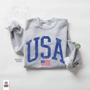 May include: A light gray sweatshirt with the word "USA" in distressed blue lettering. Below the text is a small American flag. The sweatshirt is a sport gray color.