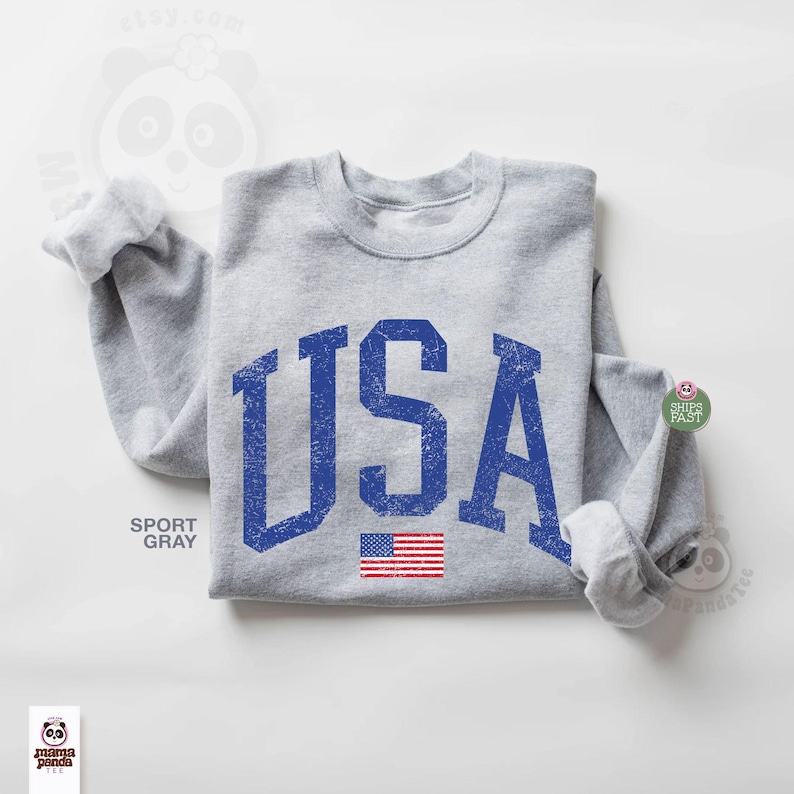 May include: A light gray sweatshirt with the word "USA" in distressed blue lettering. Below the text is a small American flag. The sweatshirt is a sport gray color.