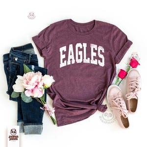 K&ouml;nnte beinhalten: Ein weinrotes T-Shirt mit dem Wort "EAGLES" in Wei&szlig;, kombiniert mit einer blauen Jeans, rosa Turnschuhen und einem Strau&szlig; rosa und wei&szlig;er Blumen. Das T-Shirt ist unten geknotet. Ein l&auml;ssiges, stilvolles Outfit.