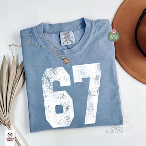 May include: A blue jean colored t-shirt with the number "67" in white, distressed lettering. The shirt has a crew neck and short sleeves. A gold necklace with a pendant is draped on the shirt. A brown hat is in the background.