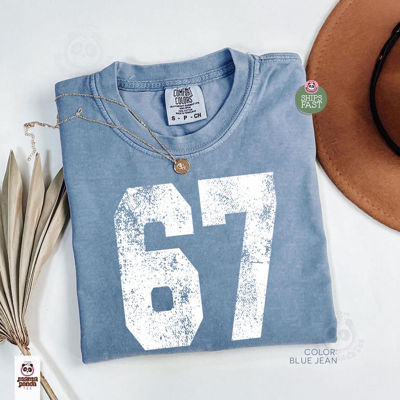May include: A blue jean colored t-shirt with the number "67" in white, distressed lettering. The shirt has a crew neck and short sleeves. A gold necklace with a pendant is draped on the shirt. A brown hat is in the background.