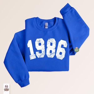 May include: A royal blue sweatshirt with the year "1986" printed in distressed white lettering across the front. The sweatshirt has a crew neck and long sleeves. A small circular graphic is on the right sleeve. The Mama Panda logo is in the lower left corner.