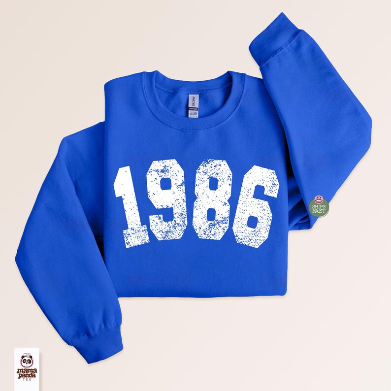 May include: A royal blue sweatshirt with the year "1986" printed in distressed white lettering across the front. The sweatshirt has a crew neck and long sleeves. A small circular graphic is on the right sleeve. The Mama Panda logo is in the lower left corner.