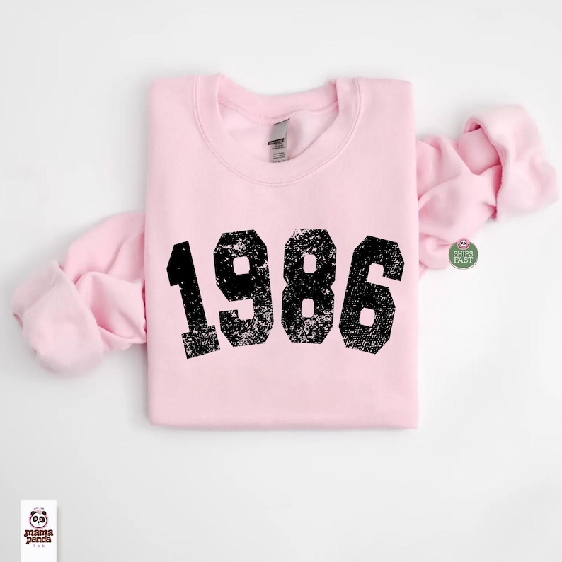 May include: A light pink sweatshirt with the year "1986" printed in distressed black lettering. The sweatshirt is folded neatly, with the sleeves draped to the side. The garment is made of a soft material.