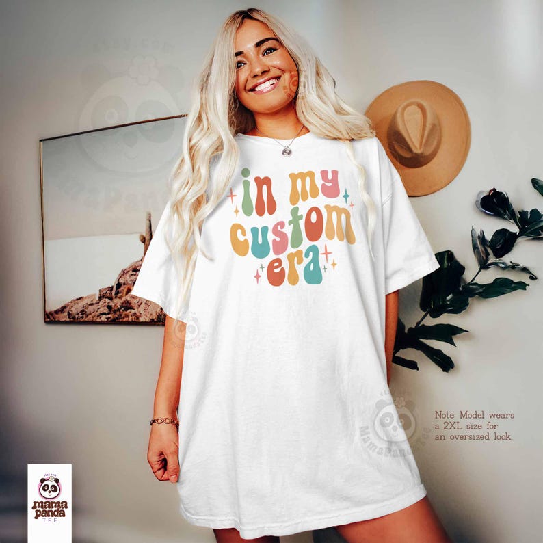 May include: White t-shirt with the text "in my custom era" in colorful retro font. The shirt has a relaxed fit. The model is wearing a 2XL size for an oversized look. A hat and artwork are in the background.