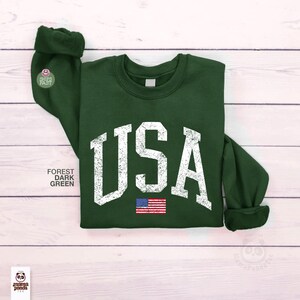 May include: Forest dark green sweatshirt with the word "USA" in distressed white lettering and a small American flag graphic. The sweatshirt has a crew neck and cuffed sleeves. The text "Forest Dark Green" is printed on the left side.