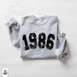 May include: A heather gray sweatshirt with the year "1986" printed in large, distressed black numbers across the front. The sweatshirt has a crew neck and long sleeves with rolled cuffs. The design is centered on the chest.