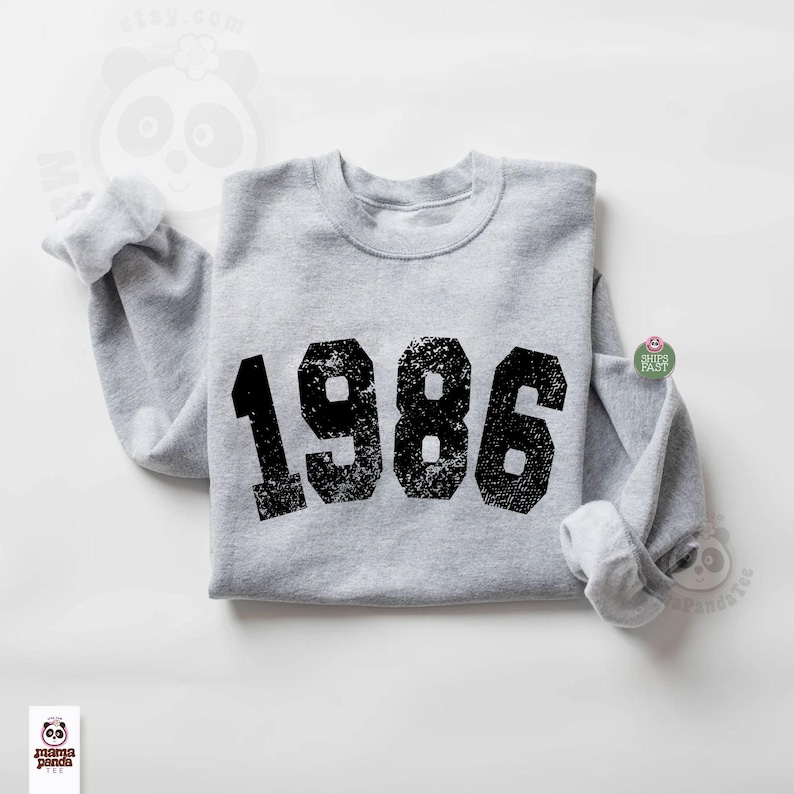 May include: A heather gray sweatshirt with the year "1986" printed in large, distressed black numbers across the front. The sweatshirt has a crew neck and long sleeves with rolled cuffs. The design is centered on the chest.