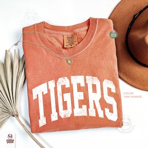 May include: A yam orange short-sleeve t-shirt featuring the word TIGERS in large, distressed white block letters across the chest. The shirt has a crew neckline with a Comfort Colors brand label and is accented by a thin gold-tone chain necklace.