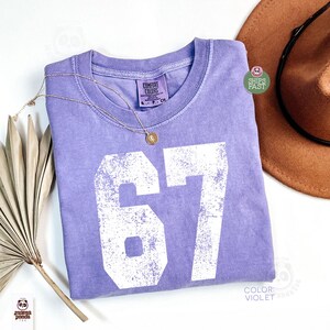 May include: A violet Comfort Colors t-shirt with the number "67" printed in white. A gold necklace with a pendant rests on the shirt. A brown hat is visible in the background.