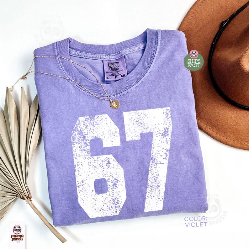 May include: A violet Comfort Colors t-shirt with the number "67" printed in white. A gold necklace with a pendant rests on the shirt. A brown hat is visible in the background.