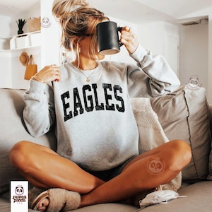 Może przedstawiać: Jasnoszara bluza z napisem "EAGLES" dużymi, wytartymi czarnymi literami. Bluza ma okrągły dekolt i długie rękawy. Czarny kubek trzymany jest przed twarzą. Osoba siedzi na szarej kanapie.
