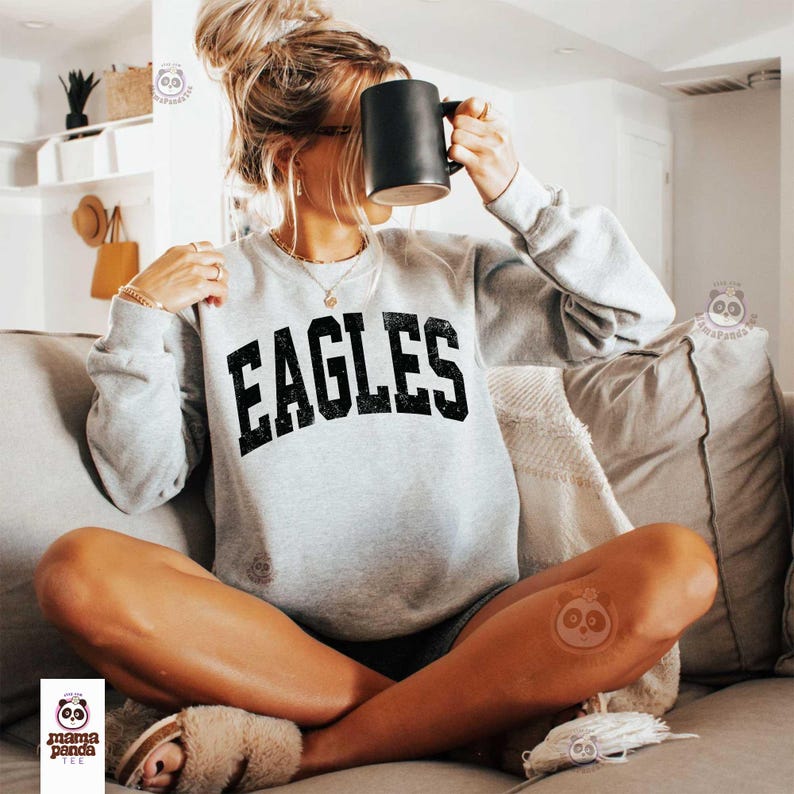 Może przedstawiać: Jasnoszara bluza z napisem "EAGLES" dużymi, wytartymi czarnymi literami. Bluza ma okrągły dekolt i długie rękawy. Czarny kubek trzymany jest przed twarzą. Osoba siedzi na szarej kanapie.