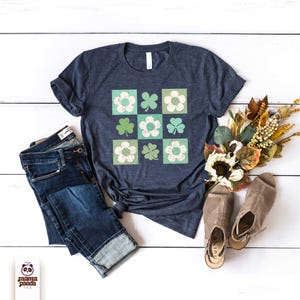Op de afbeelding: Een marineblauw t-shirt met een vintage ontwerp van groene klavers en witte bloemen in een geruit patroon. Het shirt ligt plat op een wit houten oppervlak met een blauwe jeans en bruine sandalen.