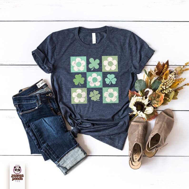 Op de afbeelding: Een marineblauw t-shirt met een vintage ontwerp van groene klavers en witte bloemen in een geruit patroon. Het shirt ligt plat op een wit houten oppervlak met een blauwe jeans en bruine sandalen.
