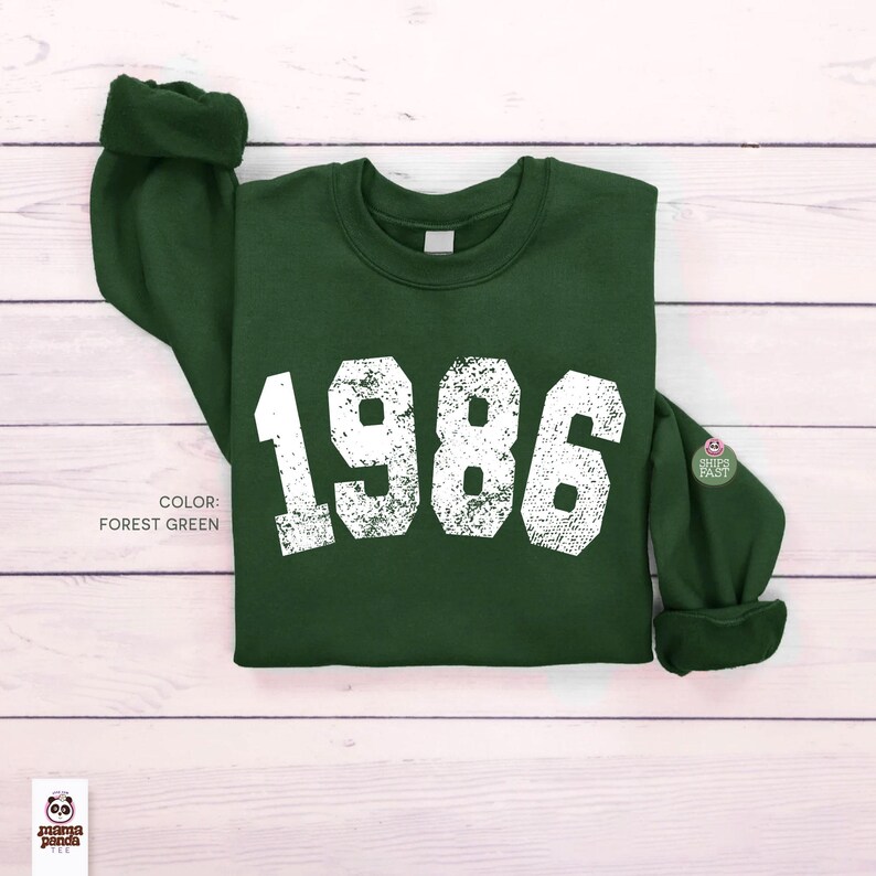 May include: Forest green sweatshirt with the year "1986" printed in distressed white lettering. The sweatshirt has a crew neck and long sleeves with cuffed ends. The color is labeled as "Forest Green".