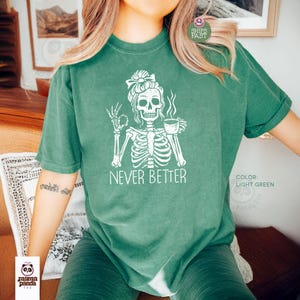 May include: A light green short-sleeve t-shirt featuring a white graphic of a skeleton with a messy bun hairstyle, holding a coffee cup and making an okay hand gesture, with the text NEVER BETTER printed in white capital letters below it.