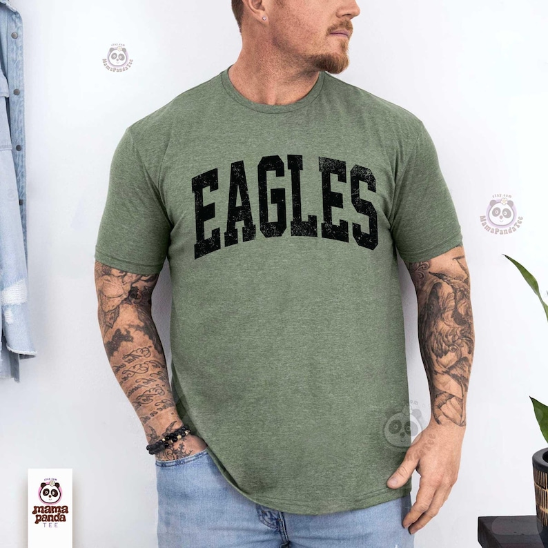 K&ouml;nnte beinhalten: Olivgr&uuml;nes T-Shirt mit dem Wort "EAGLES" in fetter, schwarzer, verwaschener Schrift. Das Shirt hat einen Rundhalsausschnitt und scheint aus einem weichen, melierten Stoff zu sein. Das Shirt wird von einer Person getragen.