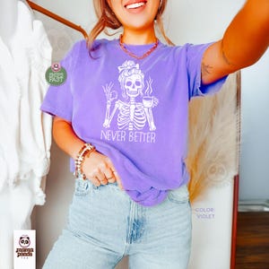 May include: A violet short-sleeve t-shirt featuring a white graphic of a skeleton with a messy bun, holding a coffee cup and making an okay hand gesture. The text NEVER BETTER is printed below the skeleton. The shirt has a crew neckline.