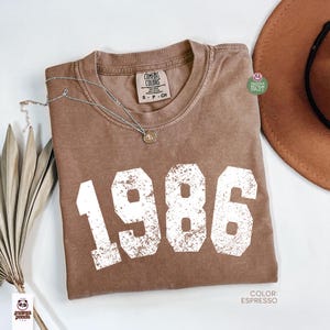 Może przedstawiać: Brązowy t-shirt w kolorze espresso z rokiem "1986" wydrukowanym na biało, w stylu vintage. Koszulka ma okrągły dekolt i kr&oacute;tkie rękawy. Srebrny naszyjnik z wisiorkiem jest na koszulce. Brązowy kapelusz jest widoczny w tle.