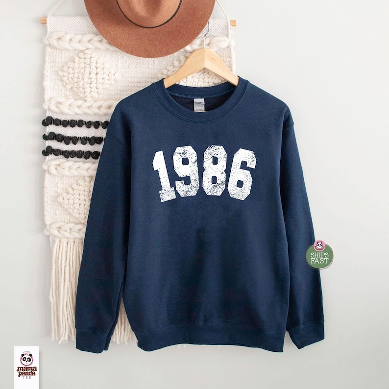 May include: Navy blue sweatshirt with the year "1986" printed in white, displayed on a wooden hanger. The sweatshirt is hanging against a neutral-toned wall with a decorative wall hanging and a brown hat.