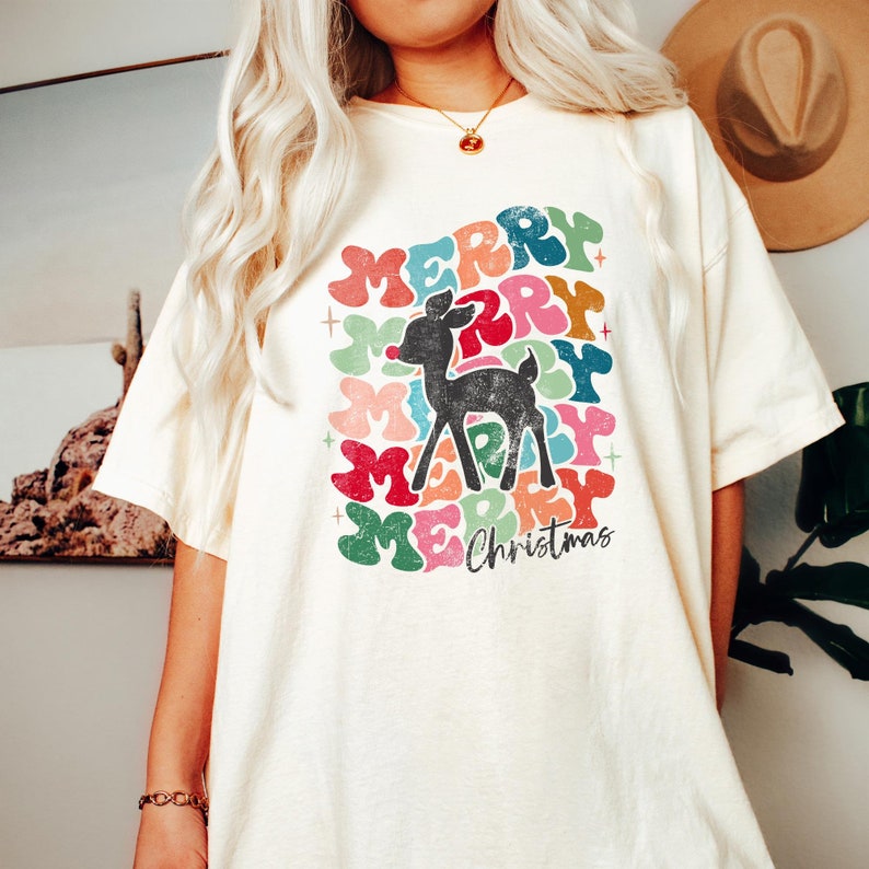 May include: A white t-shirt with a colorful graphic design of the word "Merry" repeated in a circle with a reindeer in the center. The text "Christmas" is below the circle.