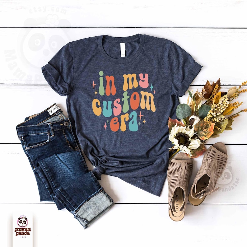 May include: A navy blue t-shirt with the text "in my custom era" in colorful retro font. The shirt is paired with blue jeans and beige sandals, arranged on a white wooden surface. A bouquet of dried flowers adds a decorative touch.