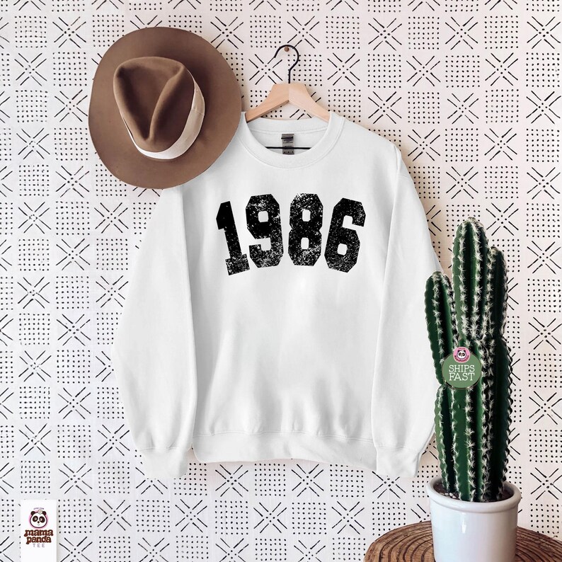 May include: A white sweatshirt with the number "1986" printed in a distressed black font. A brown fedora hat hangs on the left, and a cactus in a white pot is on the right. The background has a repeating geometric pattern.