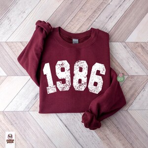 May include: A maroon sweatshirt with the year "1986" printed in distressed white lettering. The sweatshirt is made of a soft material and has a crew neck. The sleeves are folded. The background is a light wood pattern.