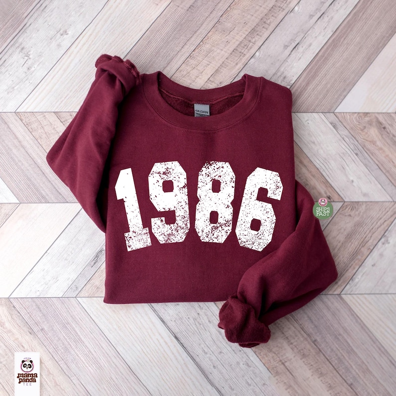 May include: A maroon sweatshirt with the year "1986" printed in distressed white lettering. The sweatshirt is made of a soft material and has a crew neck. The sleeves are folded. The background is a light wood pattern.