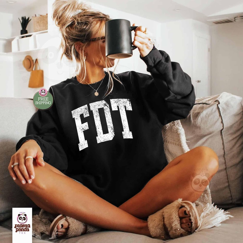 May include: Black sweatshirt with the text "FDT" in white distressed lettering. The sweatshirt is worn by a person sitting on a couch.