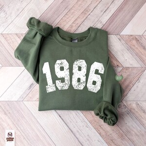 May include: An olive green sweatshirt with the year "1986" printed in distressed white lettering. The sweatshirt is folded on a light brown wooden surface. The sleeves are cuffed. The Mama Panda logo is visible in the bottom left corner.