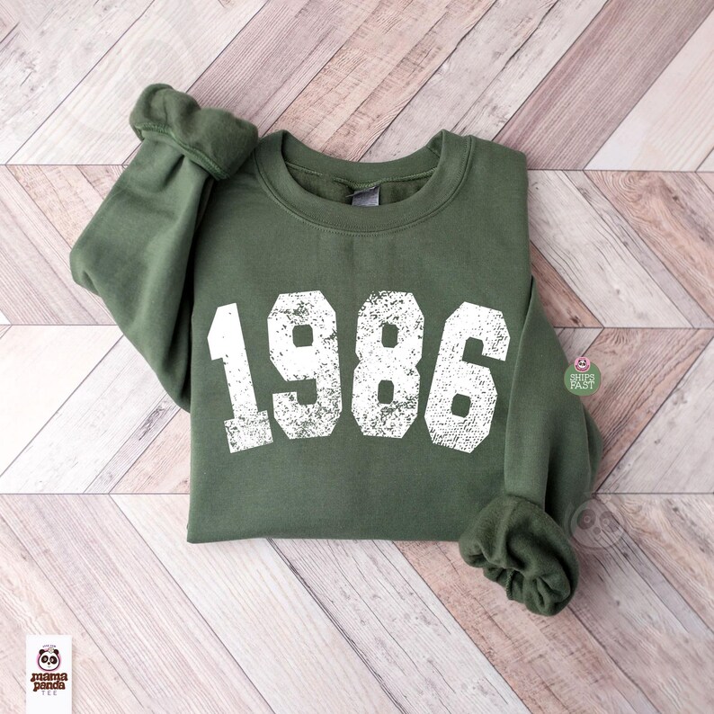 May include: An olive green sweatshirt with the year "1986" printed in distressed white lettering. The sweatshirt is folded on a light brown wooden surface. The sleeves are cuffed. The Mama Panda logo is visible in the bottom left corner.