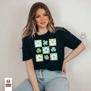 Op de afbeelding: Een zwart T-shirt met een grafische print van een geruit patroon met groene klavers en witte bloemen. Het shirt wordt gedragen door een persoon met lang bruin haar.