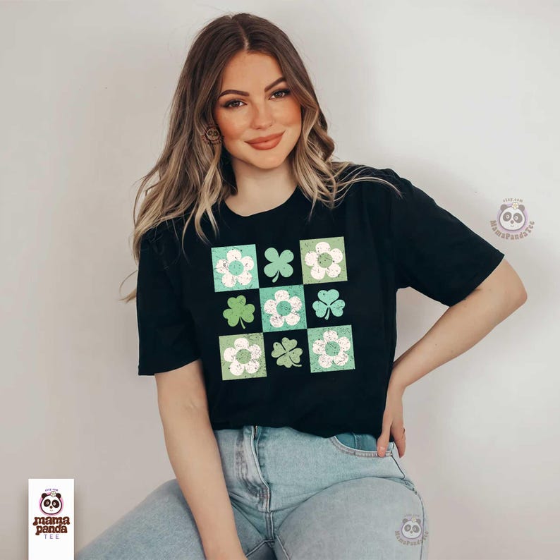 Op de afbeelding: Een zwart T-shirt met een grafische print van een geruit patroon met groene klavers en witte bloemen. Het shirt wordt gedragen door een persoon met lang bruin haar.