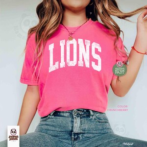 May include: A bright pink t-shirt with the word "LIONS" in white, distressed lettering. The shirt is a crew neck style and is paired with blue jeans. The color of the shirt is "Crunchberry".
