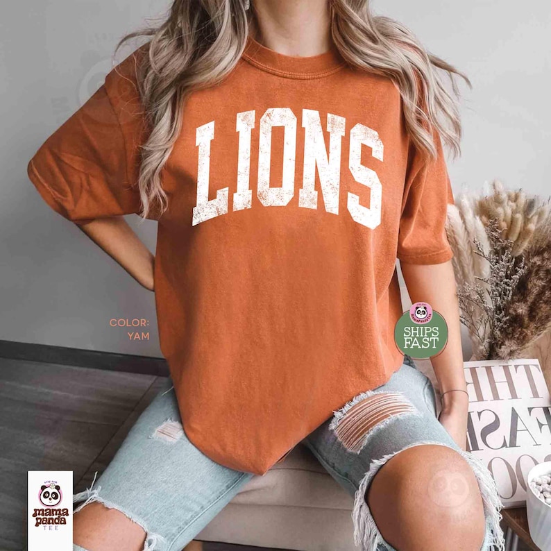 May include: A burnt orange t-shirt with the word "LIONS" in distressed white lettering. The shirt is a short-sleeved crew neck. The color is "Yam".