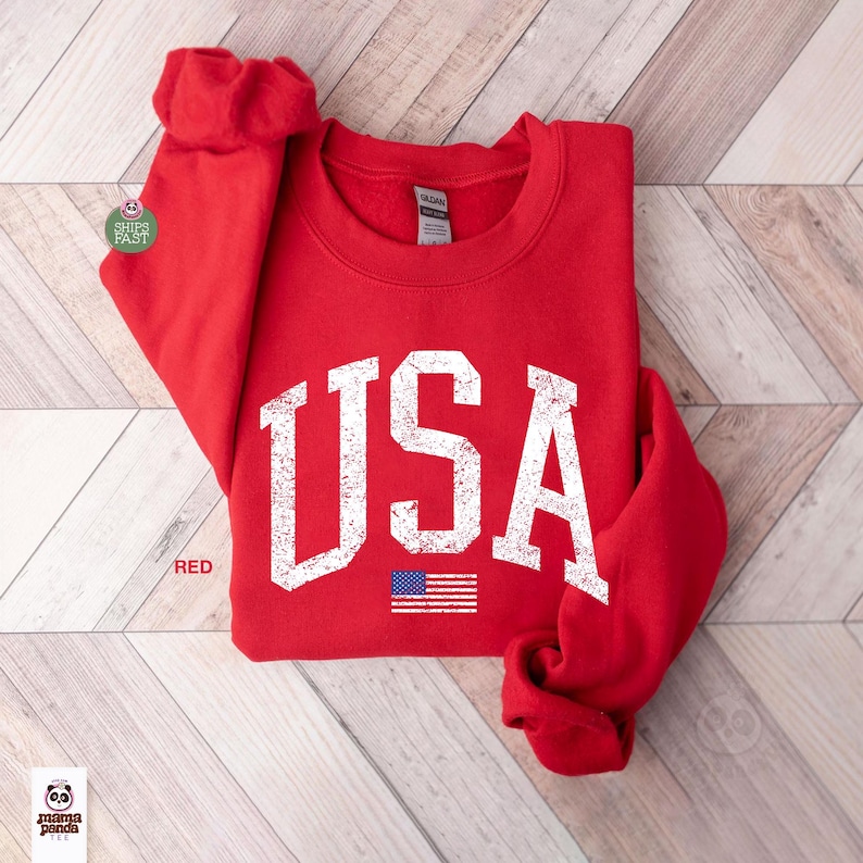 May include: A red sweatshirt with the word "USA" in distressed white lettering and a small American flag graphic. The sweatshirt is folded, with the sleeves visible. The word "RED" is printed on the bottom left.