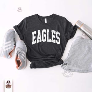 K&ouml;nnte beinhalten: Dunkelgraues T-Shirt mit dem Wort "EAGLES" in abgenutzten wei&szlig;en Buchstaben. Das Shirt wird mit grauen Shorts, Turnschuhen und einer Edelstahl-Wasserflasche kombiniert. Die Turnschuhe sind grau mit wei&szlig;en Streifen und rosa Akzenten.