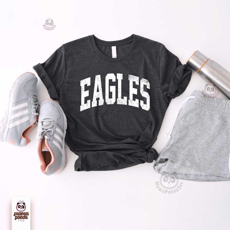 K&ouml;nnte beinhalten: Dunkelgraues T-Shirt mit dem Wort "EAGLES" in abgenutzten wei&szlig;en Buchstaben. Das Shirt wird mit grauen Shorts, Turnschuhen und einer Edelstahl-Wasserflasche kombiniert. Die Turnschuhe sind grau mit wei&szlig;en Streifen und rosa Akzenten.