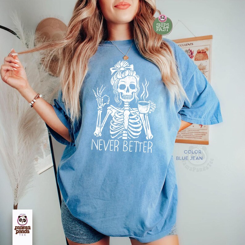 May include: A blue jean colored t-shirt featuring a white graphic of a skeleton with a messy bun hairstyle, holding a coffee cup in one hand and making an okay gesture with the other. The text "NEVER BETTER" is printed in white below the skeleton.