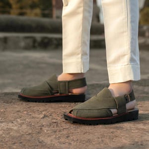 May include: A pair of olive green sandals with a buckle fastening and a thick rubber sole. The sandals are made from a canvas-like material and have a strap that goes around the ankle.