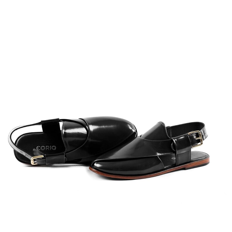 K&ouml;nnte beinhalten: Ein Paar schwarze Lackledersandalen mit spitzer Zehenpartie, Fersenriemen und einer goldfarbenen Metallschnalle. Auf der Innenseite des Fu&szlig;betts ist der Markenname CORIO in Wei&szlig; aufgedruckt. Die Sandalen haben eine flache, braune Sohle.