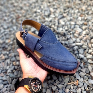 May include: A blue suede sandal with a brown leather strap and buckle. The sandal has a closed toe and a cut-out design on the side. The sandal is sitting on a bed of small grey stones.