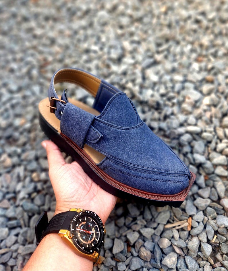 May include: A blue suede sandal with a brown leather strap and buckle. The sandal has a closed toe and a cut-out design on the side. The sandal is sitting on a bed of small grey stones.