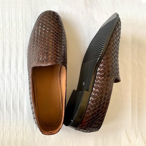 May include: A pair of dark brown woven leather loafers with a slip-on design. One shoe is positioned to show the interior tan lining, while the other is flipped to reveal the black ridged rubber sole with the number 12 printed on the heel area.