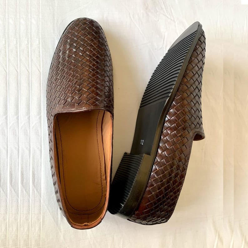 May include: A pair of dark brown woven leather loafers with a slip-on design. One shoe is positioned to show the interior tan lining, while the other is flipped to reveal the black ridged rubber sole with the number 12 printed on the heel area.