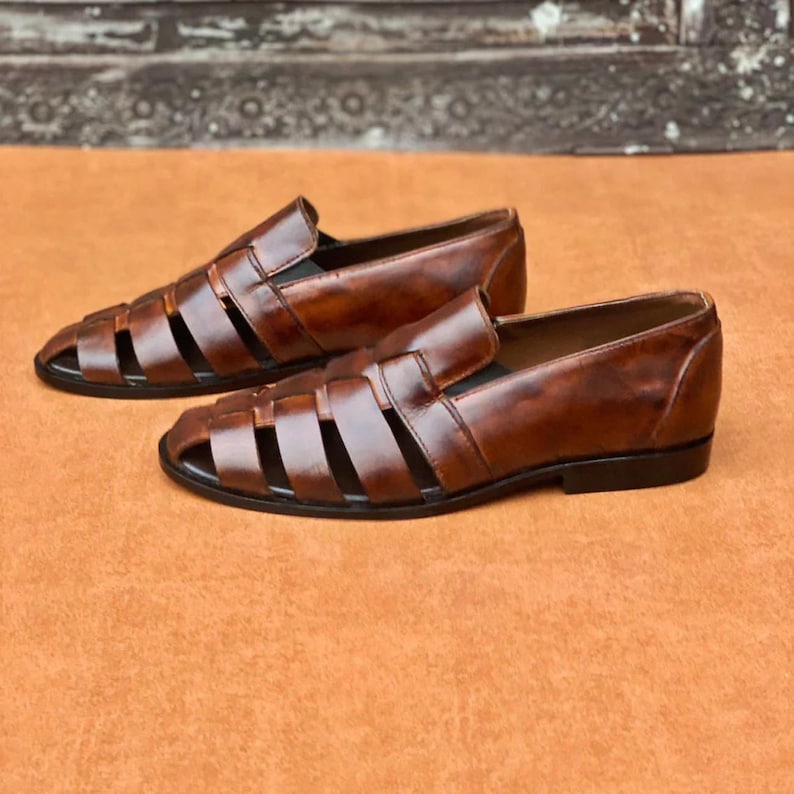 K&ouml;nnte beinhalten: Ein Paar braune Leder-Loafer im Huarache-Stil mit einem geflochtenen Riemendesign &uuml;ber dem Vorderblatt, einer geschlossenen Fersenkappe und einer flachen schwarzen Sohle. Das Leder hat ein poliertes, patiniertes Finish mit sichtbaren Farbnuancen.